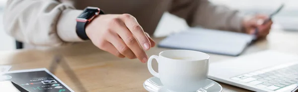 Vista recortada del hombre de negocios tomando taza de café cerca de la computadora portátil y papel en la oficina, pancarta - foto de stock