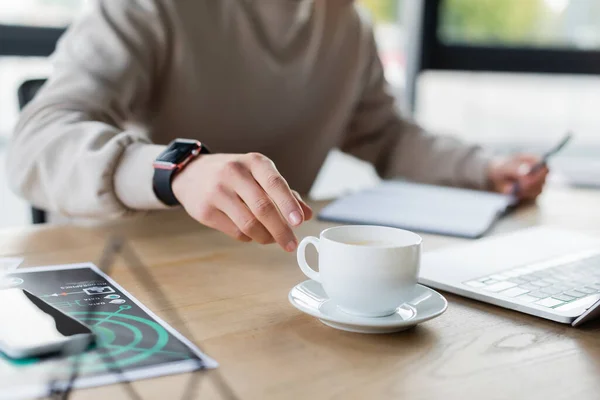 Обрезанный вид бизнесмена, берущего чашку кофе рядом с устройствами и ноутбуком в офисе — стоковое фото