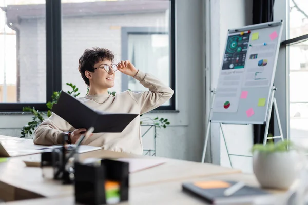 Smiling transgender person holding eyeglasses and paper folder while working in office — Stock Photo