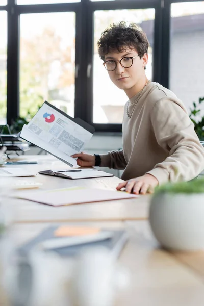 Молодий бізнесмен тримає папери з розмовами біля записника і ноутбука в офісі. — стокове фото