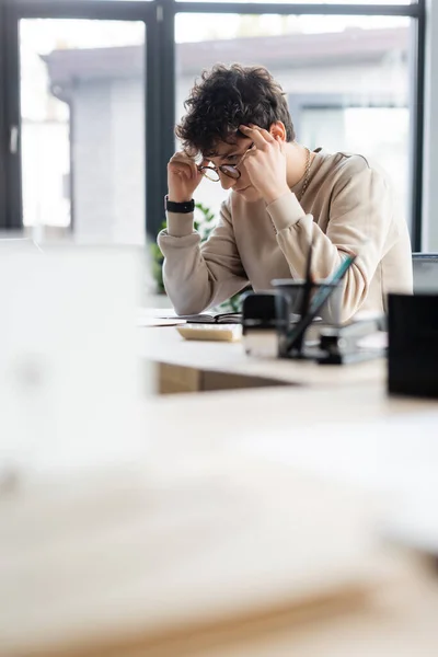 Uomo d'affari pensieroso in occhiali seduto vicino notebook e calcolatrice offuscata in ufficio — Foto stock