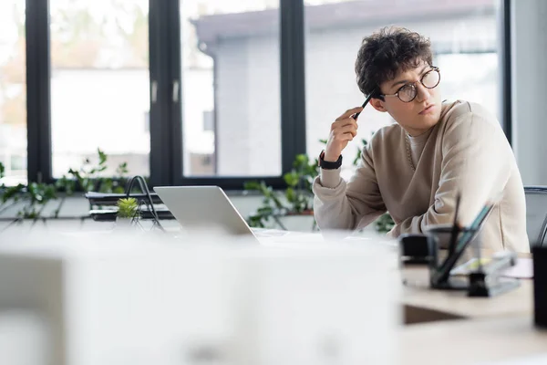 Pensativo empresario sosteniendo pluma cerca de la computadora portátil en la oficina - foto de stock