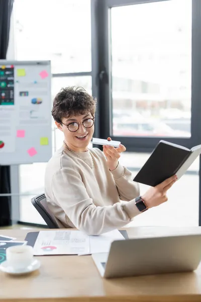 Positive businessman recording voice message on smartphone and holding notebook near laptop in office — Stock Photo