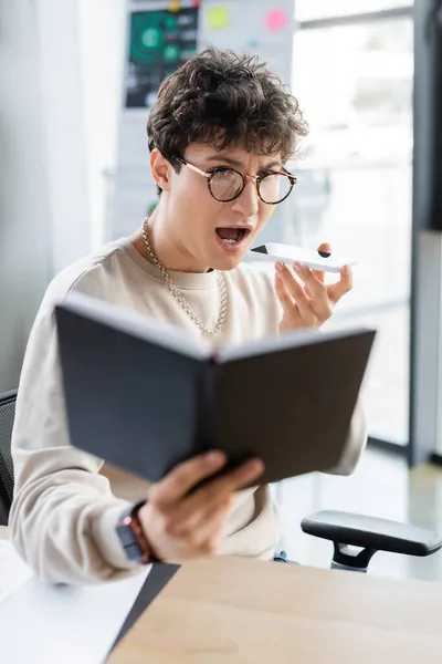 Astonished businessman recording voice message on smartphone and looking at blurred notebook in office — Stock Photo