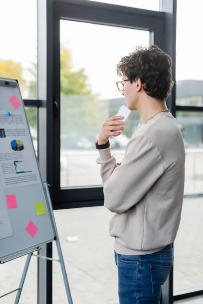 Vista laterale del giovane uomo d'affari che tiene lo smartphone mentre guarda il flip chart in ufficio — Foto stock