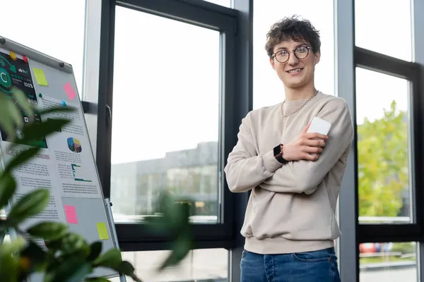 Hombre de negocios sonriente sosteniendo el teléfono inteligente y mirando la cámara cerca del rotafolio y la planta en la oficina - foto de stock