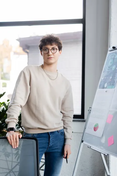 Junger Transgender blickt im Büro in die Kamera neben Stuhl und Flipchart — Stockfoto