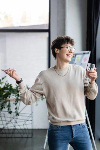 Uomo d'affari sorridente in auricolare con smartphone e bicchiere d'acqua in ufficio — Foto stock