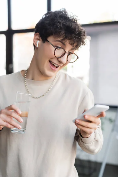 Positive transgender person in earphone using smartphone and holding glass of water in office — Stock Photo