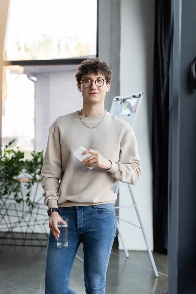 Молодой трансгендер в наушниках держит стакан воды и смартфон в офисе — стоковое фото
