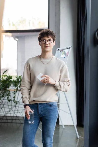 Young transgender person in earphones holding glass of water and smartphone in office — Stock Photo