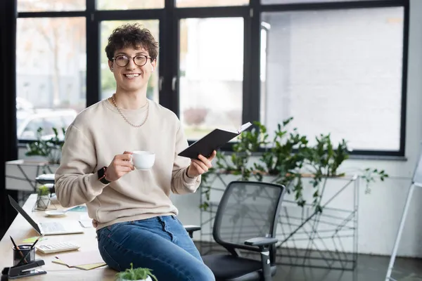 Positiver Geschäftsmann mit Kopfhörer, Kaffee und Notizbuch in der Hand, während er im Büro in die Kamera schaut — Stockfoto