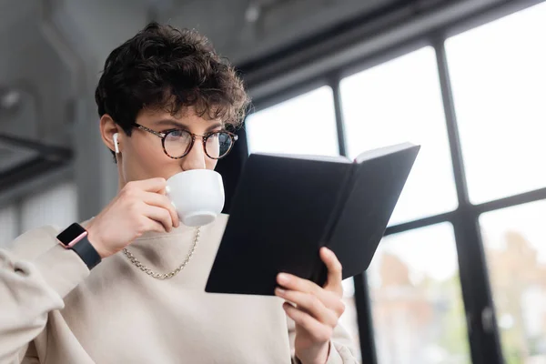 Молодой бизнесмен в наушниках пьет кофе и смотрит на блокнот в офисе — стоковое фото