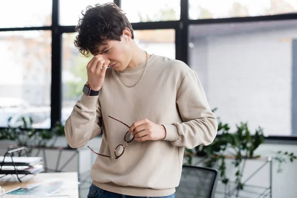 Uomo d'affari stanco in auricolare che tiene gli occhiali in ufficio — Foto stock