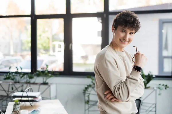 Young transgender person in earphone holding eyeglasses and smiling at camera in office — Stock Photo