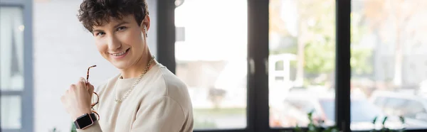 Positive transgender person in earphone holding eyeglasses in office, banner — Stock Photo