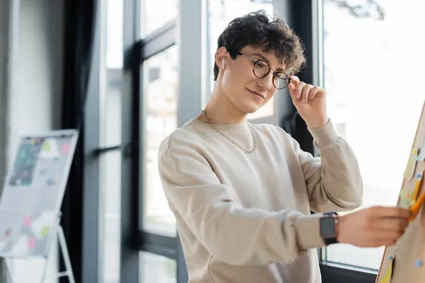 Sonriente persona transgénero en gafas y auriculares de pie cerca de la junta con notas adhesivas en la oficina - foto de stock