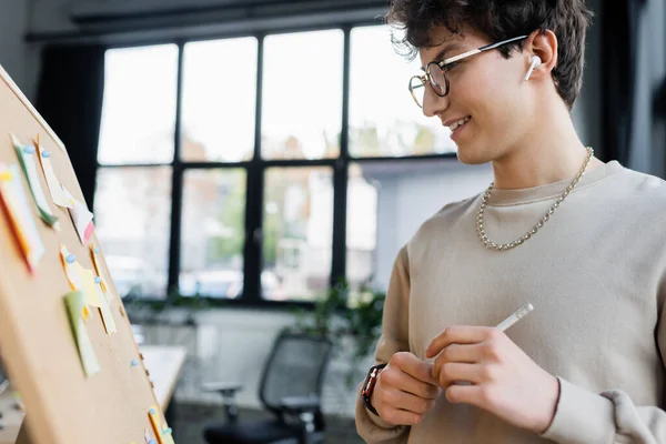 Young transgender person in earphone looking at board with sticky notes in office — Stock Photo