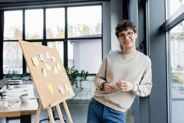 Transgender person in earphones holding pencil near board with sticky notes in office — Stock Photo