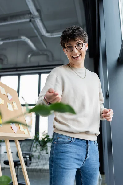 Positive transgender person in earphones looking at camera near board with sticky notes in office — Stock Photo