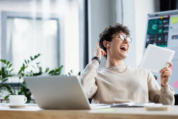 Uomo d'affari allegro in auricolare che tiene tablet digitale vicino a computer portatile e caffè in ufficio — Foto stock