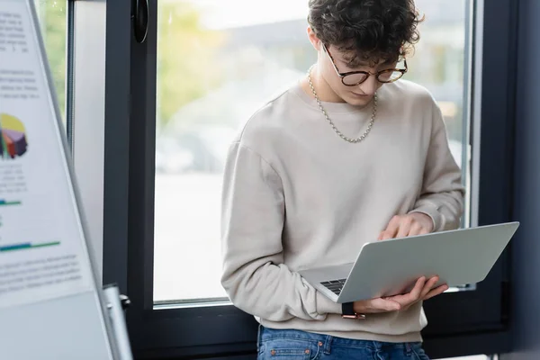 Jovem empresário em óculos usando laptop perto gráfico flip borrado no escritório — Fotografia de Stock