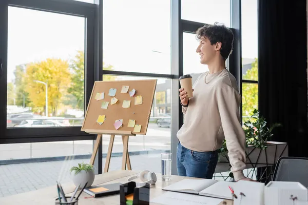 Vista lateral de una persona transexual sonriente sosteniendo café para ir cerca de la mesa de trabajo y el tablero con notas adhesivas en la oficina - foto de stock