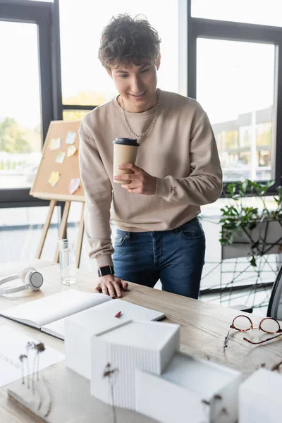 Empresário positivo segurando copo de papel e olhando para o modelo de construção perto notebook no escritório — Fotografia de Stock