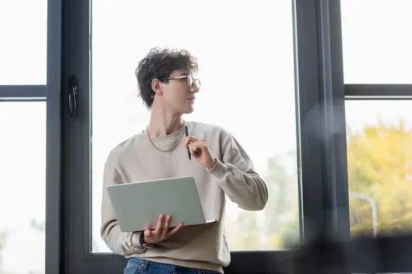 Transgender person in eyeglasses holding laptop and pen near window in office — Stock Photo
