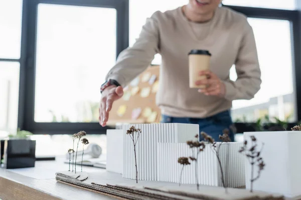 Cropped view of blurred businessman holding paper cup and pointing at model of building in office — Stock Photo