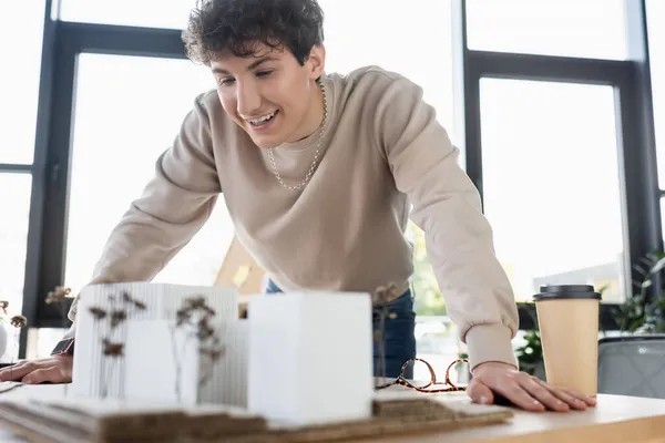 Hombre de negocios alegre mirando modelo de edificio cerca de anteojos y café para ir en oficina - foto de stock