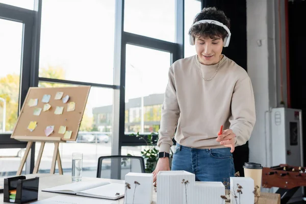 Geschäftsmann mit Kopfhörer hält Stift in der Nähe Modell des Gebäudes und Getränke im Büro — Stockfoto