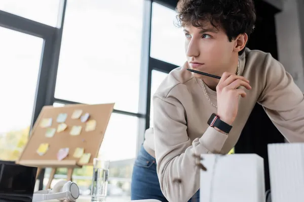 Homme d'affaires pensif tenant un stylo près du modèle flou du bâtiment et des écouteurs sur la table dans le bureau — Photo de stock