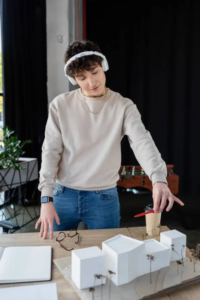 Молода трансгендерна людина в навушниках тримає ручку біля моделі будівлі та кави, щоб піти в офіс — стокове фото