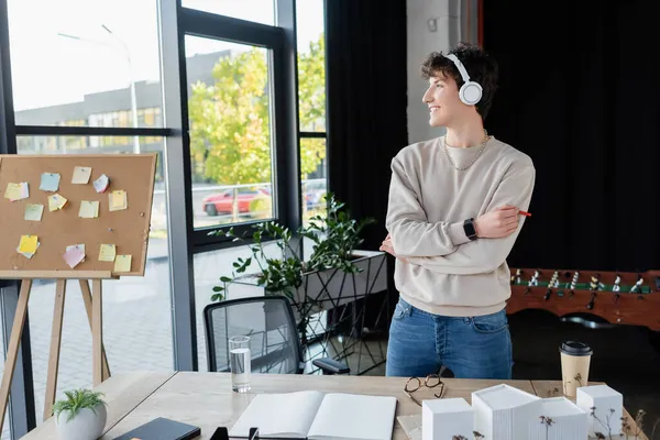 Vue latérale de l'homme d'affaires souriant dans les écouteurs debout près de la table de travail dans le bureau — Photo de stock