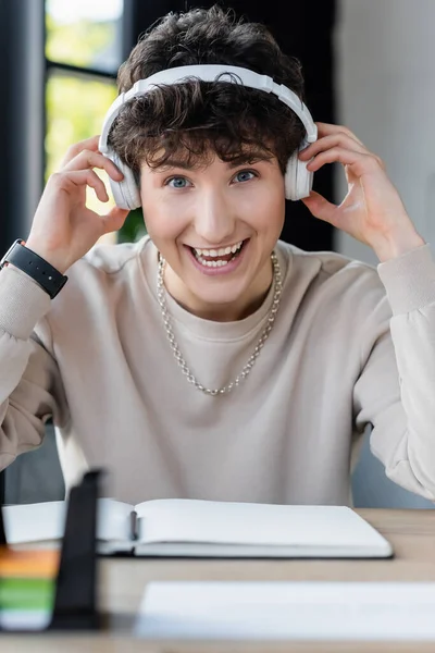 Positive transgender person in headphones looking at camera near blurred notebook in office — Stock Photo