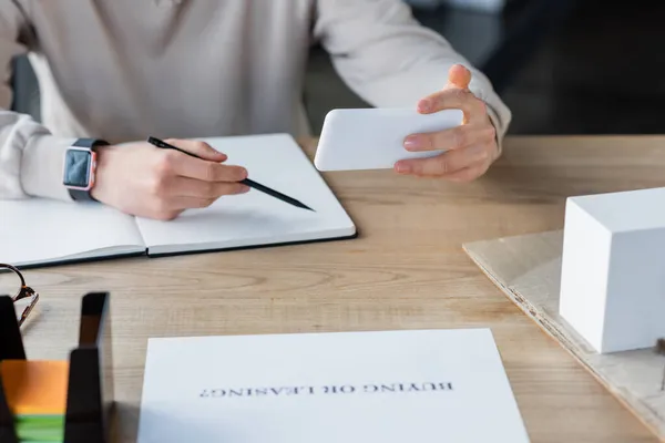 Cropped view of businessman holding smartphone while writing on notebook near paper with buying or leasing lettering in office — Stock Photo