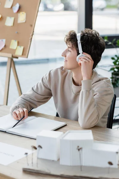 Side view of young transgender person in headphones sitting near notebook and model of house in office — Stock Photo