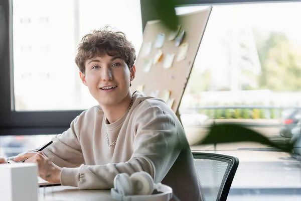 Positive transgender person looking at camera near blurred headphones in office — Stock Photo