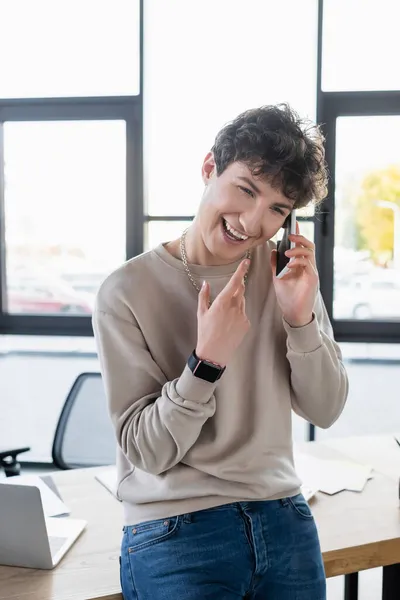 Gai transgenre personne parlant sur smartphone près d'un ordinateur portable sur la table dans le bureau — Photo de stock