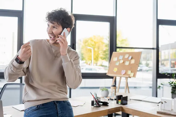 Persona transgénero positiva hablando en smartphone cerca de mesa de trabajo en la oficina - foto de stock