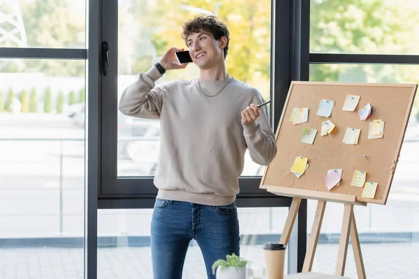 Young businessman talking on smartphone near board with sticky notes in office — Stock Photo