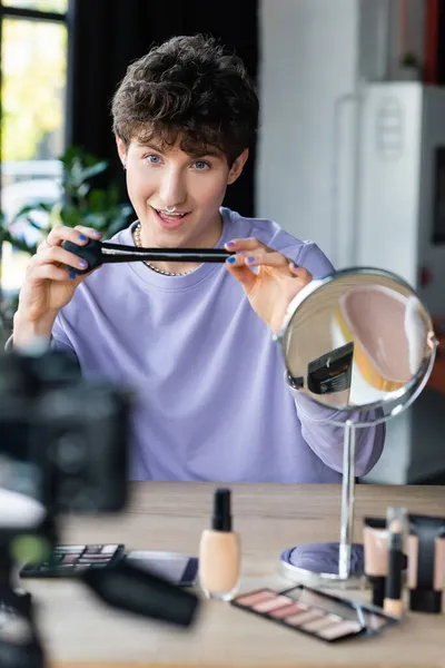 Artista de maquiagem transexual sorridente segurando escova cosmética perto de cosméticos e câmera digital borrada — Fotografia de Stock