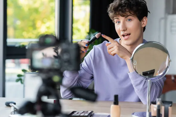 Artista de maquillaje transgénero apuntando al cepillo cosmético cerca del espejo y la cámara digital - foto de stock