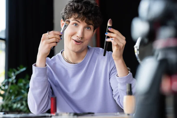 Sonriente persona transgénero sosteniendo cepillos cosméticos cerca de la base de la cara borrosa y la cámara digital - foto de stock