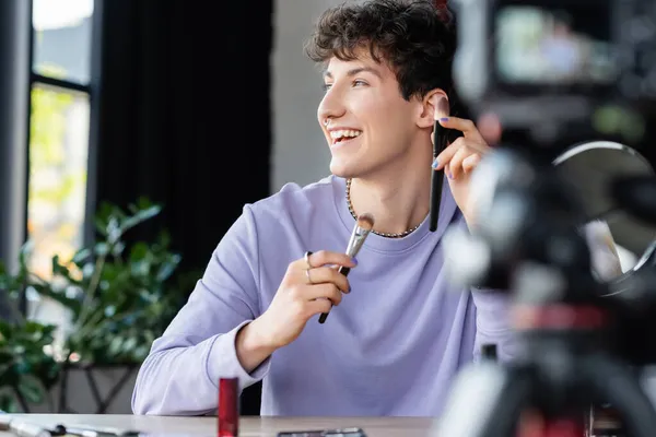 Positive transgender person holding cosmetic brushes near blurred digital camera — Stock Photo