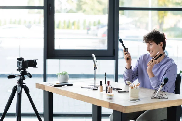 Allegro artista del trucco transgender che tiene pennelli cosmetici vicino ai cosmetici e alla fotocamera digitale — Foto stock