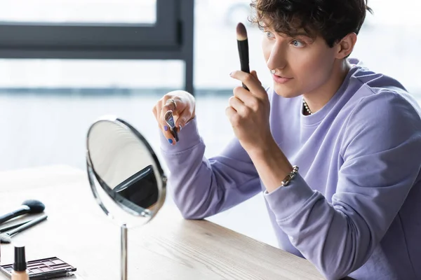 Jovem artista de maquiagem transexual segurando escovas cosméticas perto do espelho — Fotografia de Stock