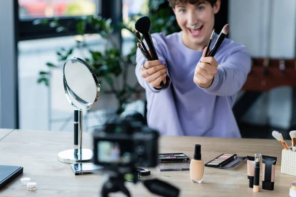 Maquillaje transgénero borroso artista que sostiene pinceles cosméticos cerca de la cámara digital en el estudio - foto de stock
