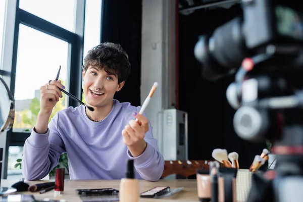 Happy transgender blogger holding cosmetic brushes near blurred digital camera — Stock Photo